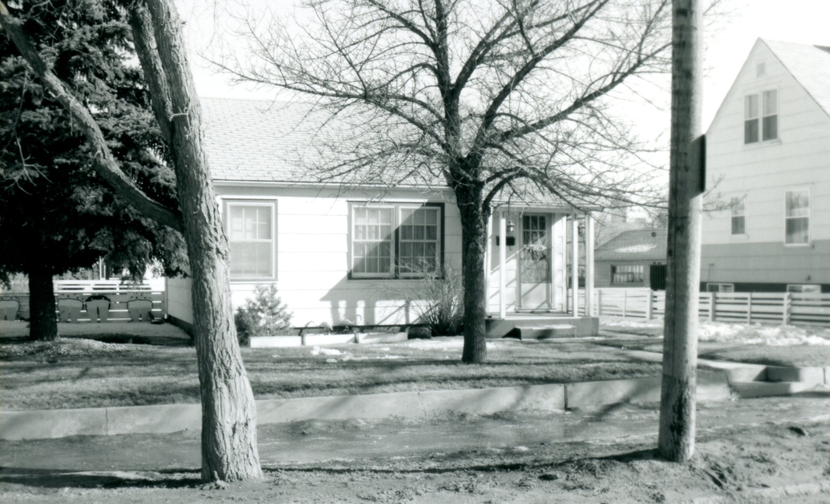 A modest single family house facing a street with sidewalk, trees, and power pole in front.